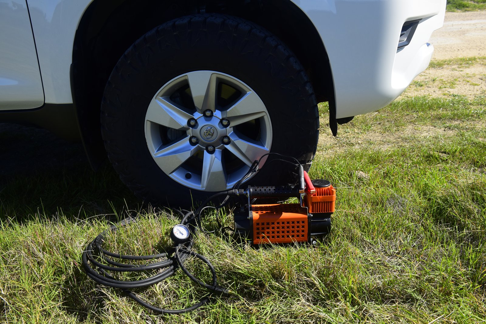 Vehicle and Camping Gear Orange Maxi Trac 12V air compressor on grass next to the Prado wheel, with coiled inflation hose and pressure gauge