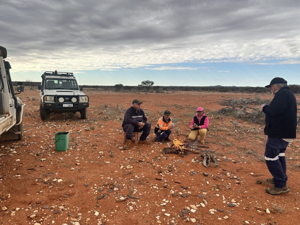 Four people around a small campfire on quartz-scattered red dirt in the WA goldfields