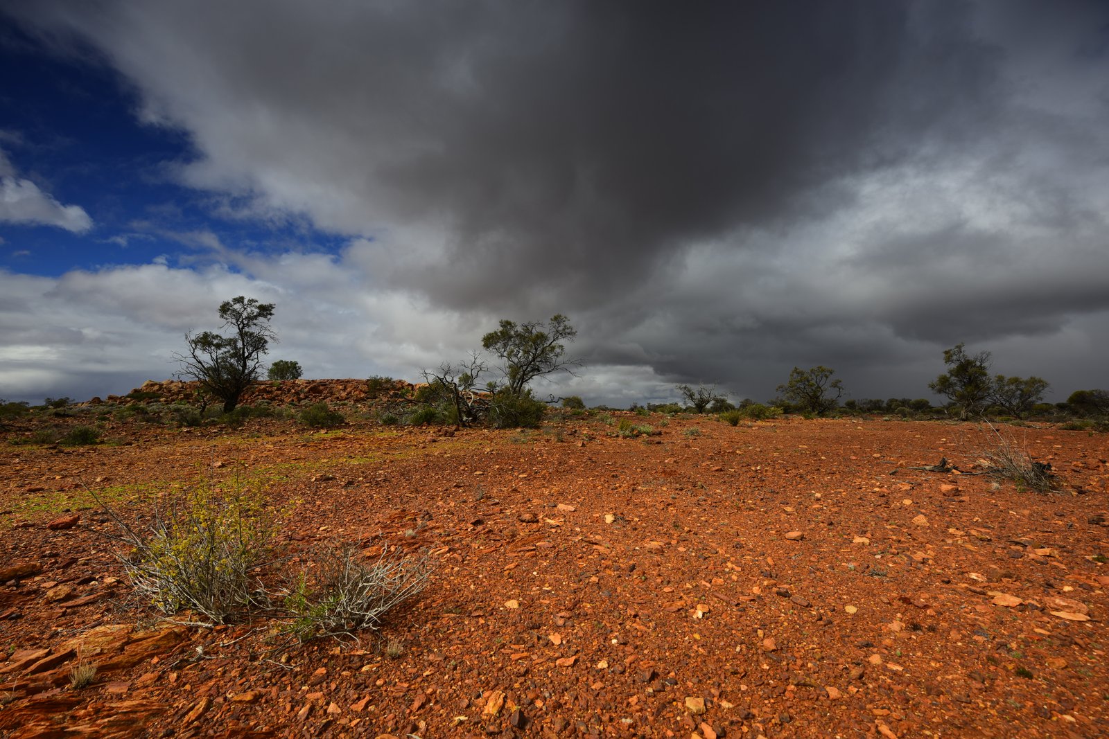 A heavy grey storm front building over a low ironstone rise and red gravel ground.