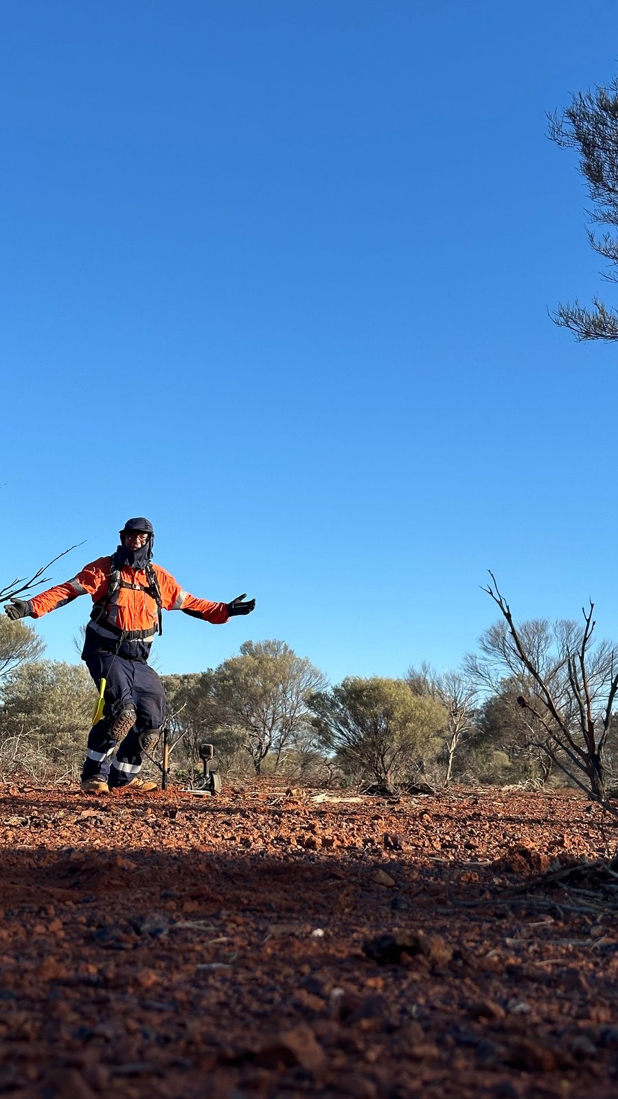 Rob in hi-vis, a neck-flap cap and knee pads, arms spread wide on red stony ground, the metal detector on the ground behind him.