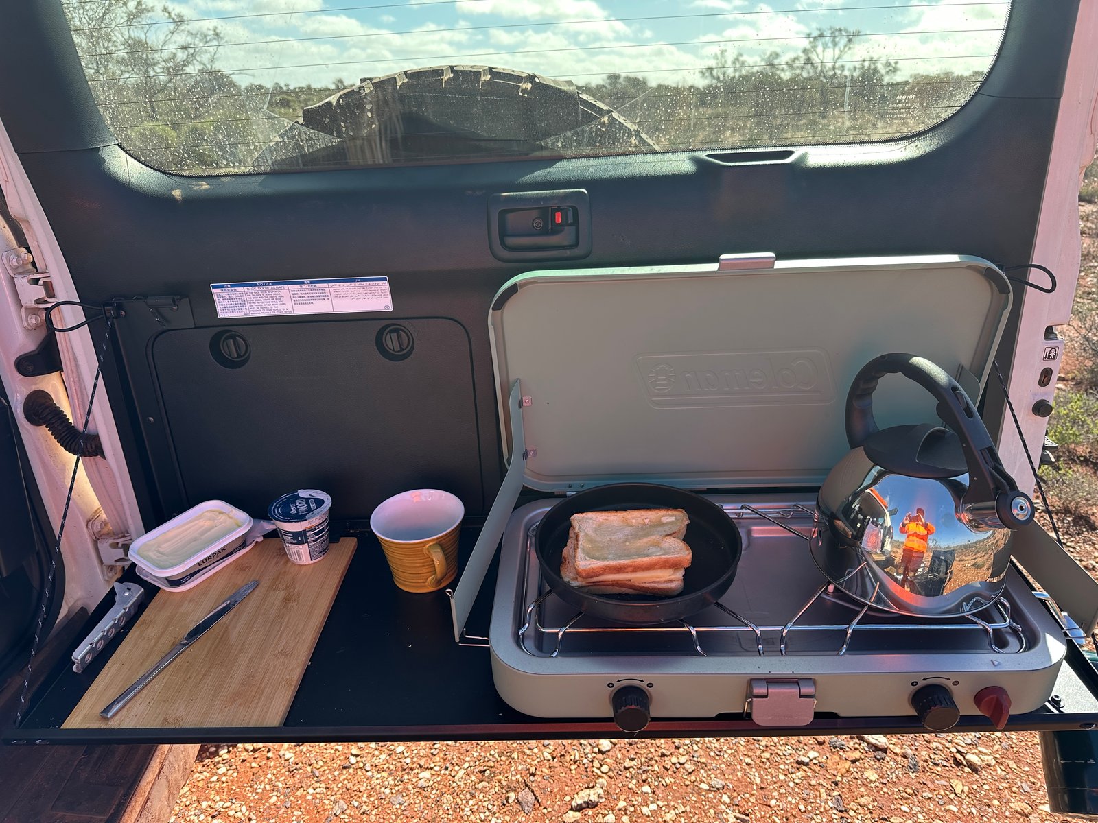 Did I Mention the Junk? A toasted cheese sandwich in a frying pan on a camp stove on the back of a four-wheel drive, with the photographer in hi-vis reflected in the kettle.