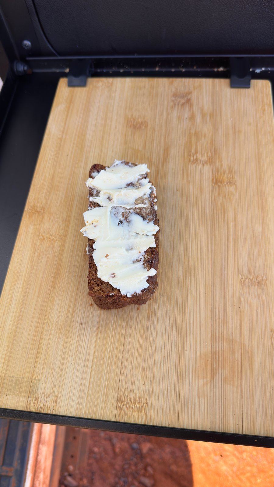 A thick slice of buttered date loaf on a bamboo chopping board at the back of the Prado, red dirt below.