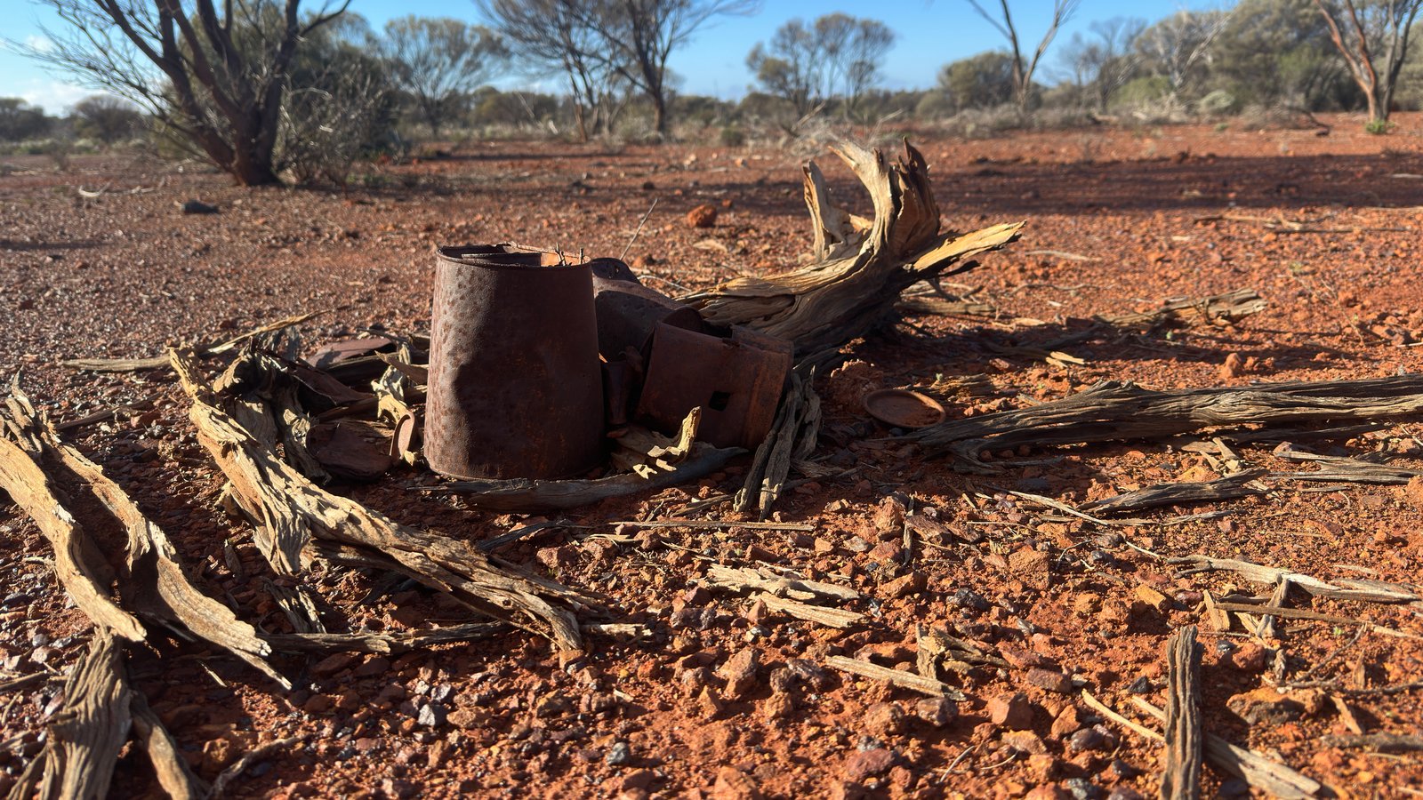 Old rusted tins lying among dead, sun-bleached timber on red stony ground.