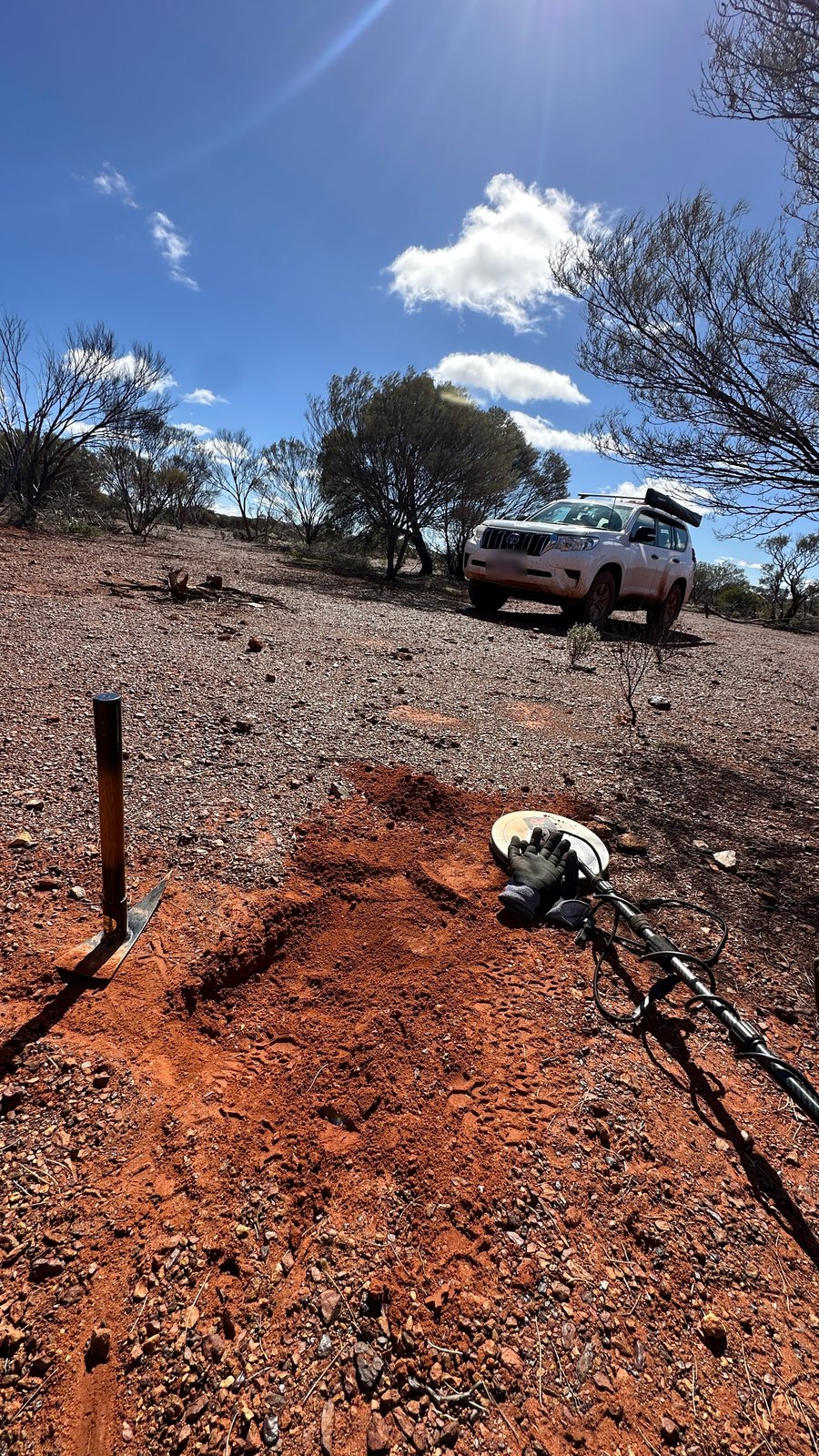 A freshly dug hole in red dirt with a pick, gloves and metal detector beside it, and a white Prado parked a short way behind.