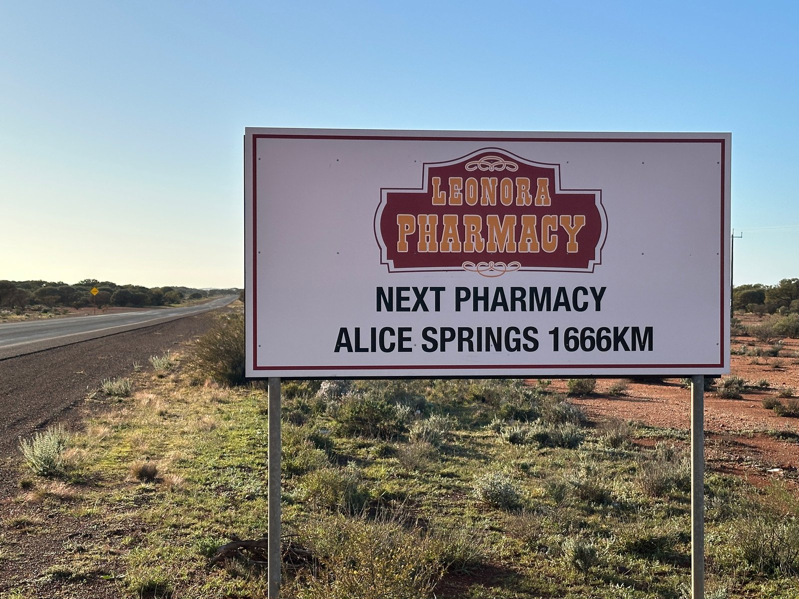 Did I Mention the Junk? Roadside sign for the Leonora Pharmacy reading "Next pharmacy Alice Springs 1666 km", beside an empty highway.