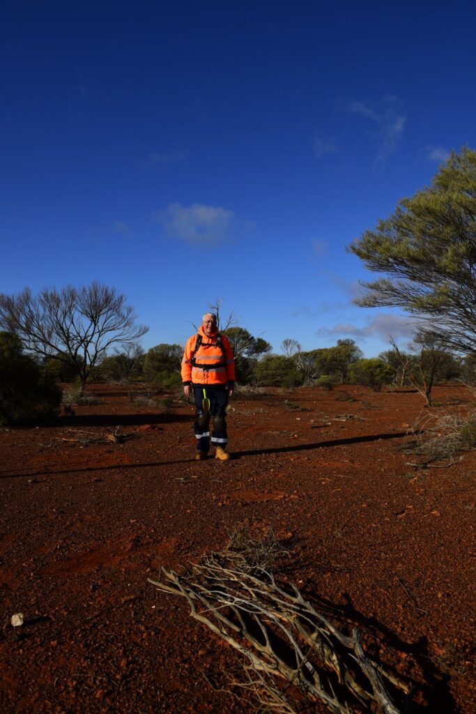 Rob in a hi-vis jacket standing still on red dirt under a deep blue sky, with scrub and trees behind him.