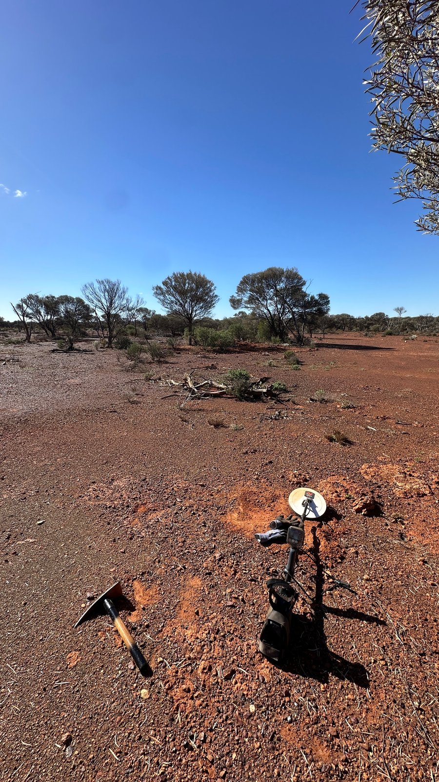 A metal detector and a pick laid down beside a small dig on open red ground under a clear blue sky.